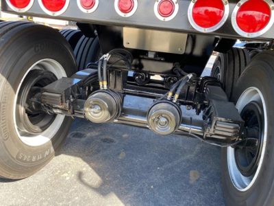 Trailer rear view showing suspension and dual axles with leaf springs, axles, and tires. Visible components include the axle housing, air hoses, and mounting brackets under the dump trailer.