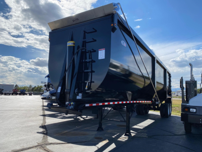 Dump trailer in a parking lot with a raised front, side markings not legible, and a hitch area visible. The trailer is dark blue/black with reflective red-white stripe along the bottom edge.