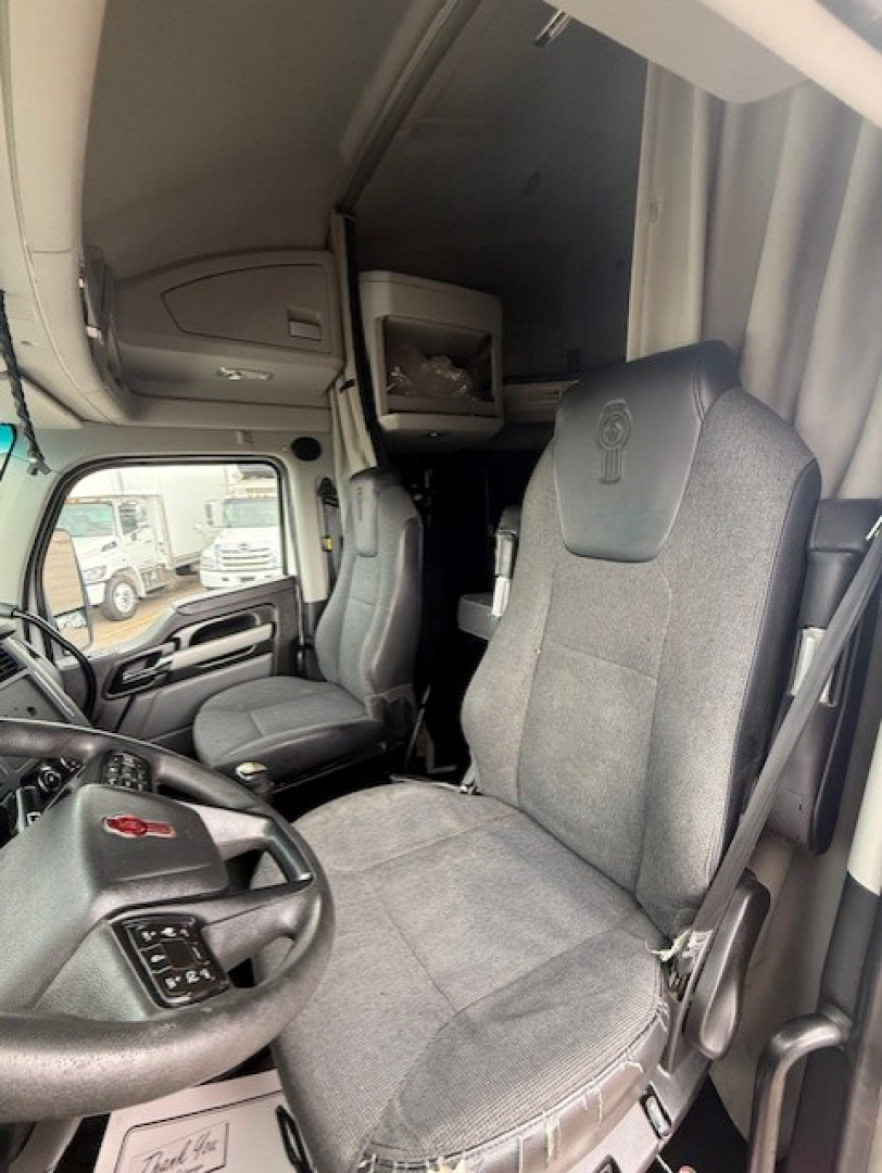 Cabin interior of a 2023 Kenworth T680 showing driver seat and passenger seat, overhead sleeper cabinet, steering wheel, and dashboard.