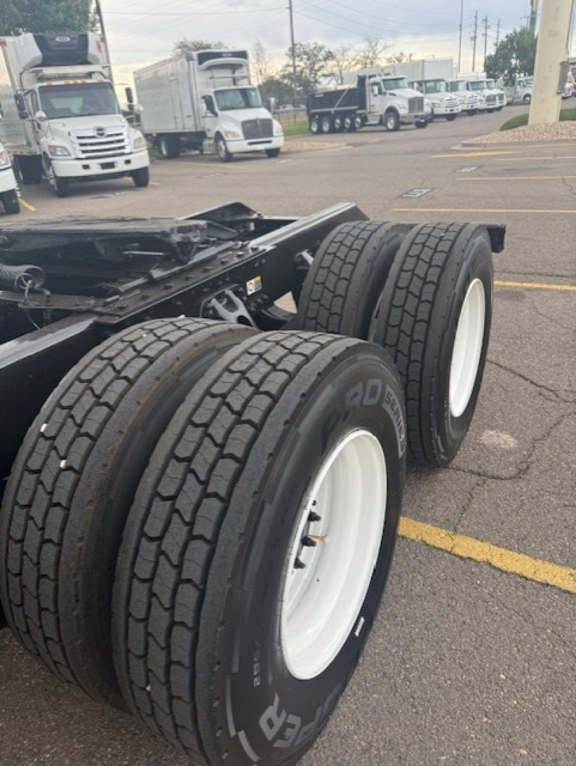 Dual rear truck axles with large dual tires and white rims on a Kenworth tractor chassis; background shows a truck lot with parked sleeper cabs.