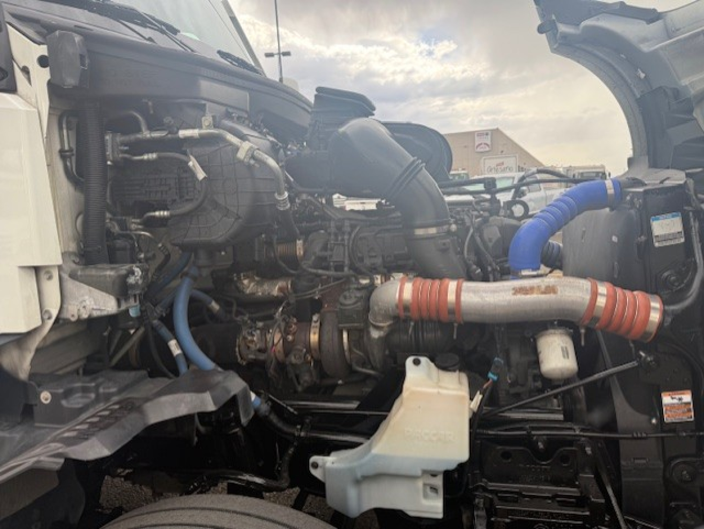 Kenworth T680 engine bay showing large turbocharger and blue and red hoses with coolant reservoir and various engine components.