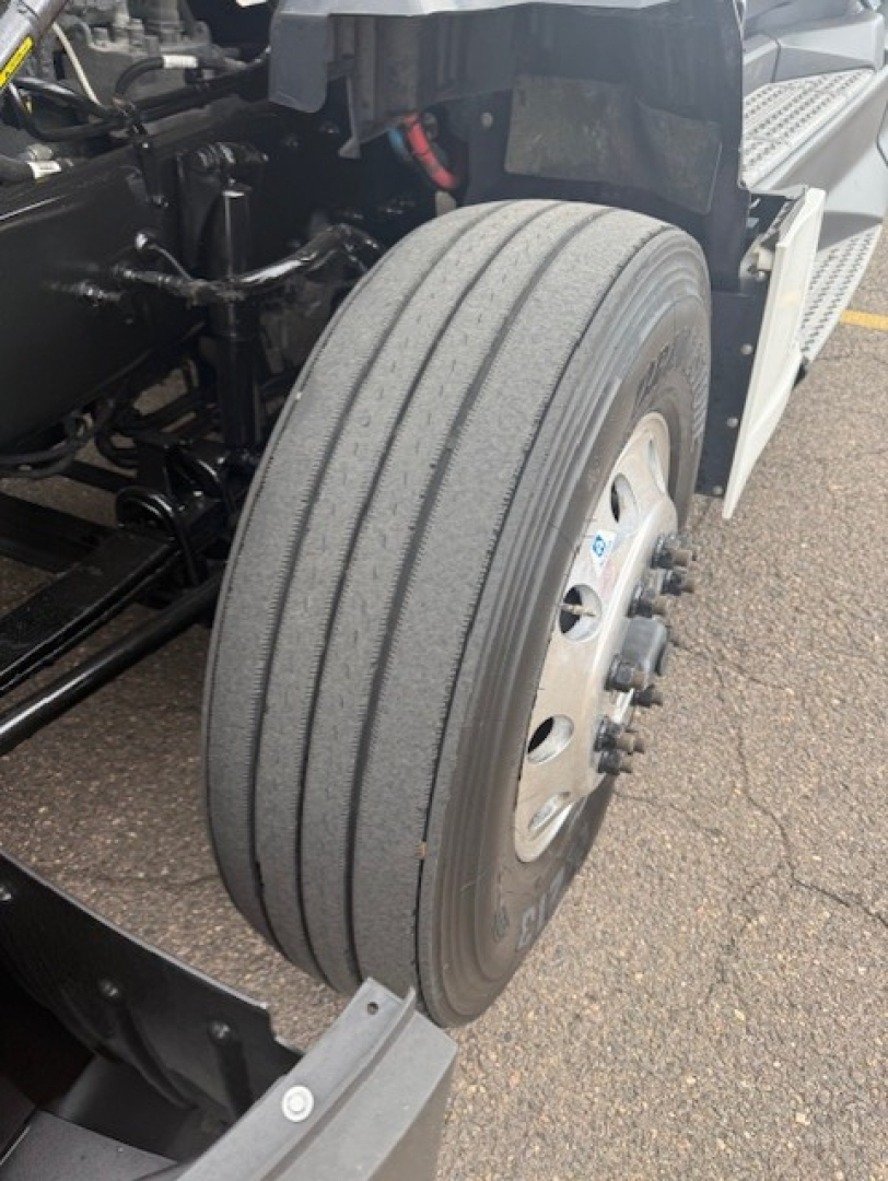 Kenworth T680 front wheel and suspension view showing front axle with tire and wheel, partial engine bay components, and chassis members.