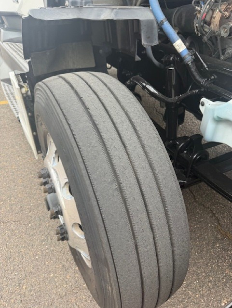 Front wheel and suspension of a Kenworth T680 with large dual rear tire and exposed chassis; visible components include blue hose, black metal brackets, and a light blue reservoir cap.