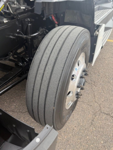 Kenworth T680 front wheel and suspension view showing front axle with tire and wheel, partial engine bay components, and chassis members.