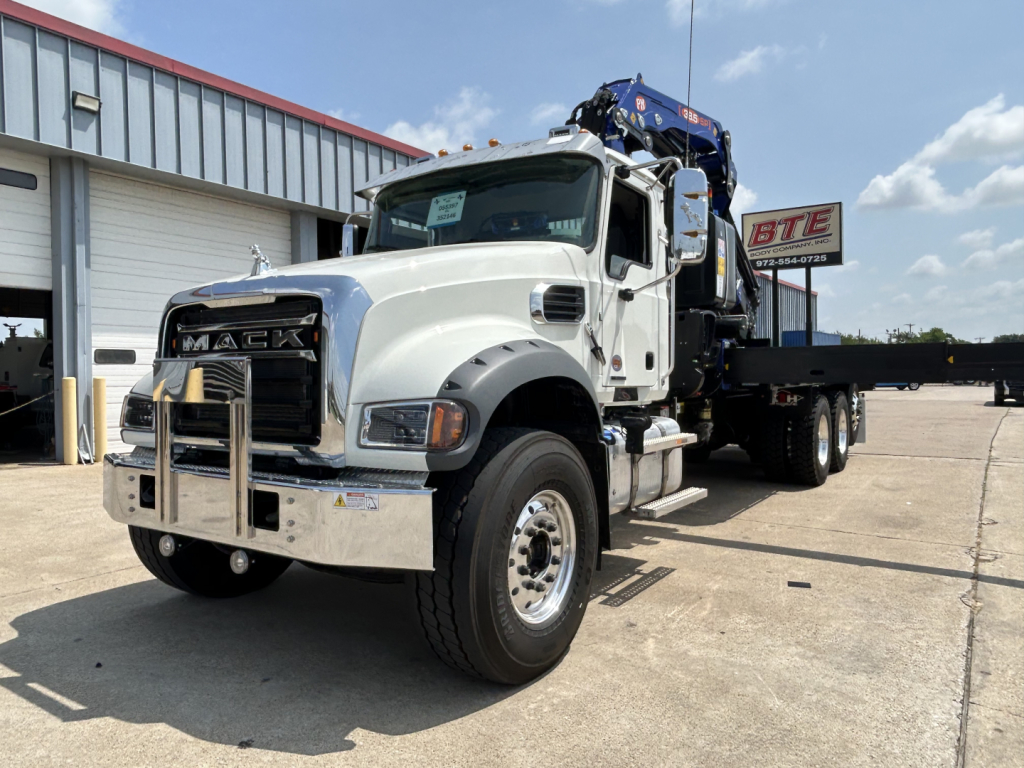 2027 Mack GR64F crane truck showing front grille and bumper with winch; chrome bumper guard; cab with Mack logo; truck bed with crane in rear.