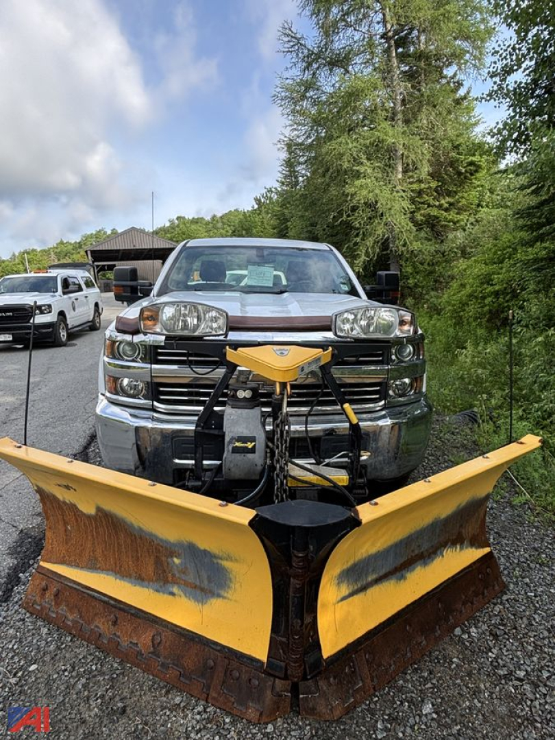 Chevrolet Silverado 3500HD pickup with a large yellow snow plow attachment mounted to the front, chrome grille, 4x4 truck, outdoor lot with trees and other vehicles.