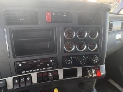 Kenworth T680 dashboard: cluster gauges and controls; left panel shows radio unit, multiple switches; top center red hazard button; HVAC controls and gauges visible.