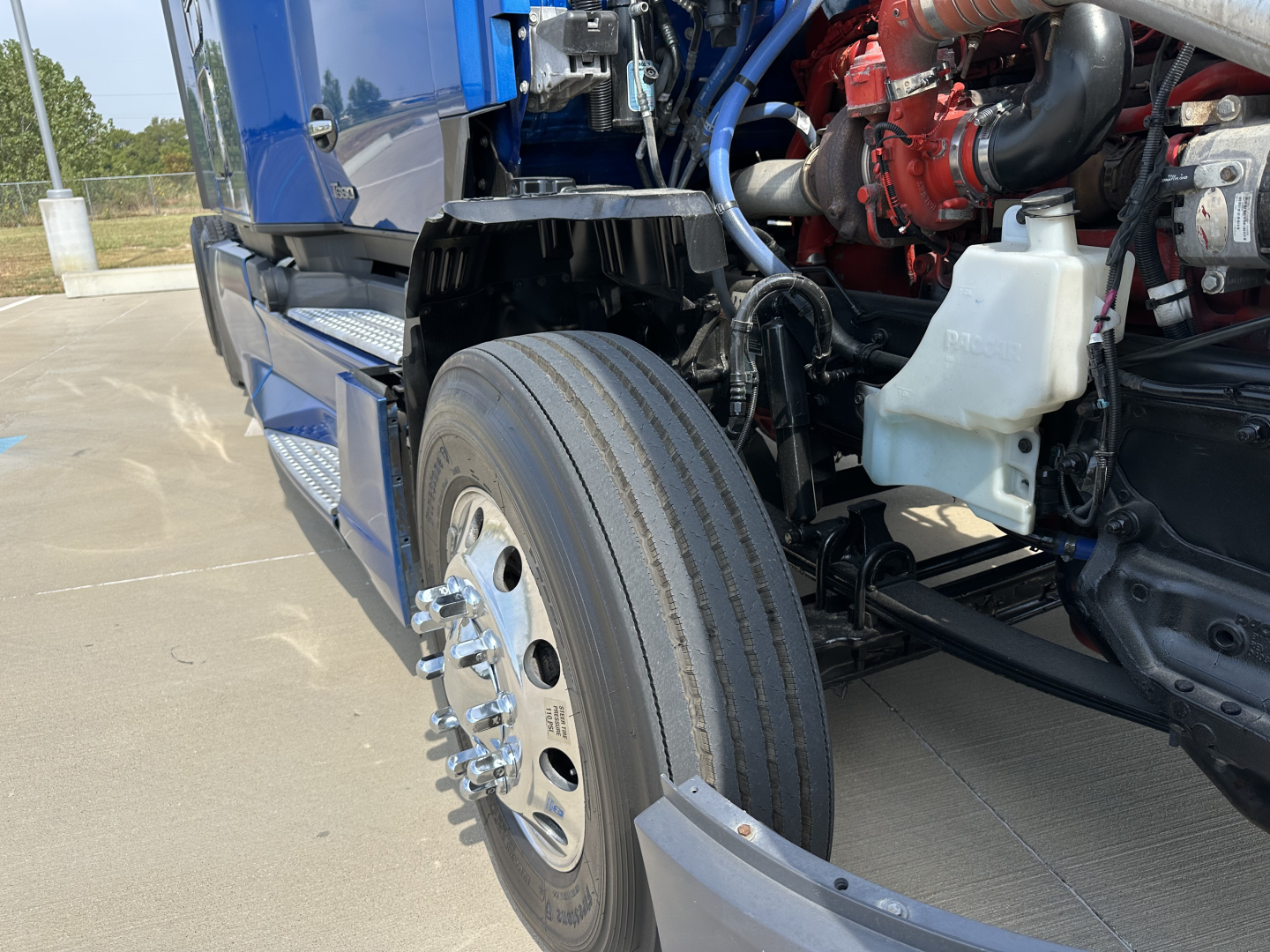 Kenworth T680 front wheel area showing front tire, wheel hub, and engine bay with red turbo piping and white coolant reservoir.
