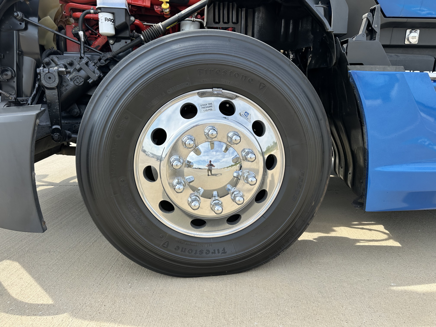 Kenworth T680 front wheel and chrome hub showing tire and wheel assembly, engine bay and blue fender in background.