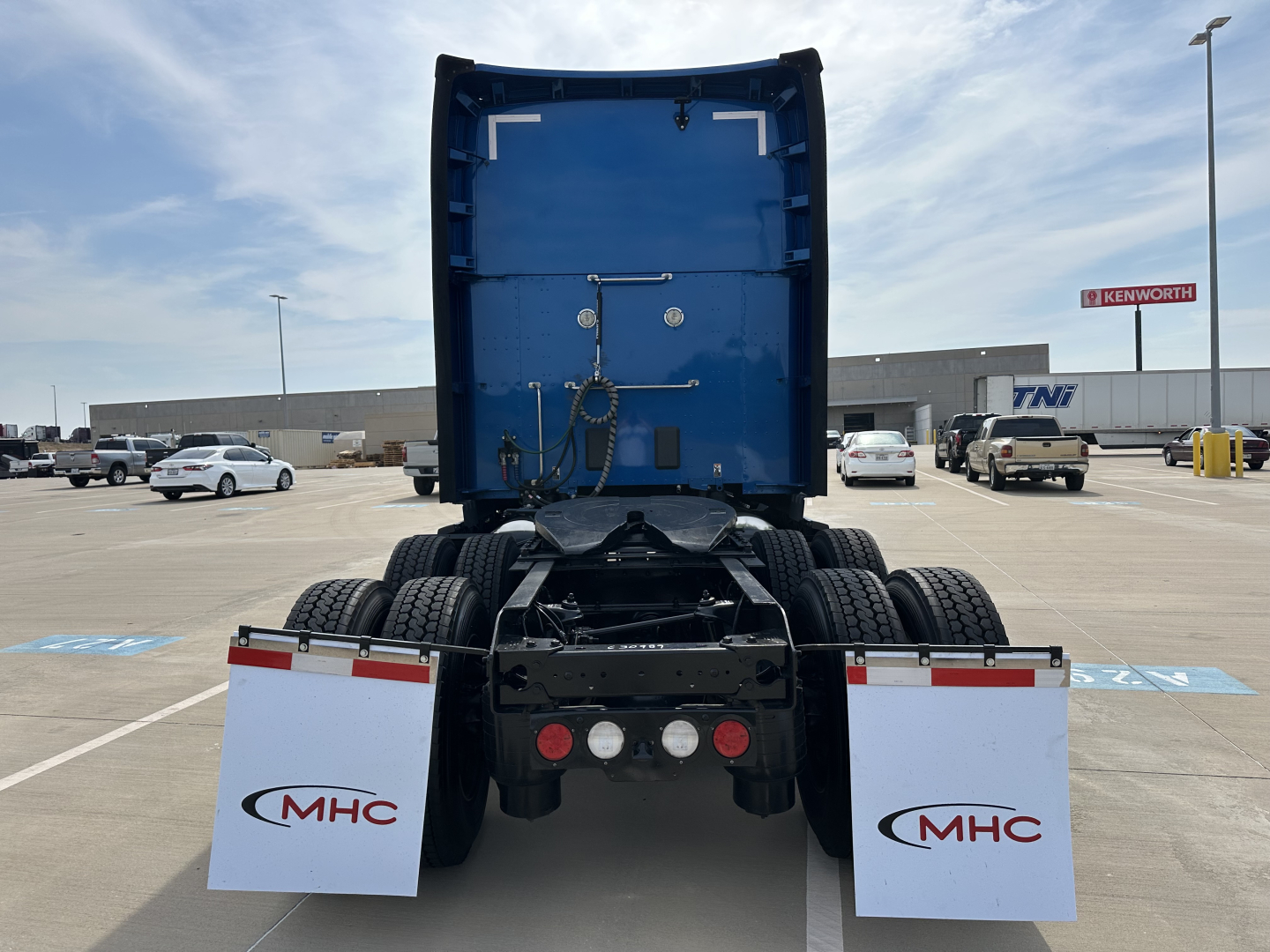 Kenworth T680 rear view showing chassis and dual rear axles with mud flaps and MHC branding, blue cab interior visible through open sleeper area.