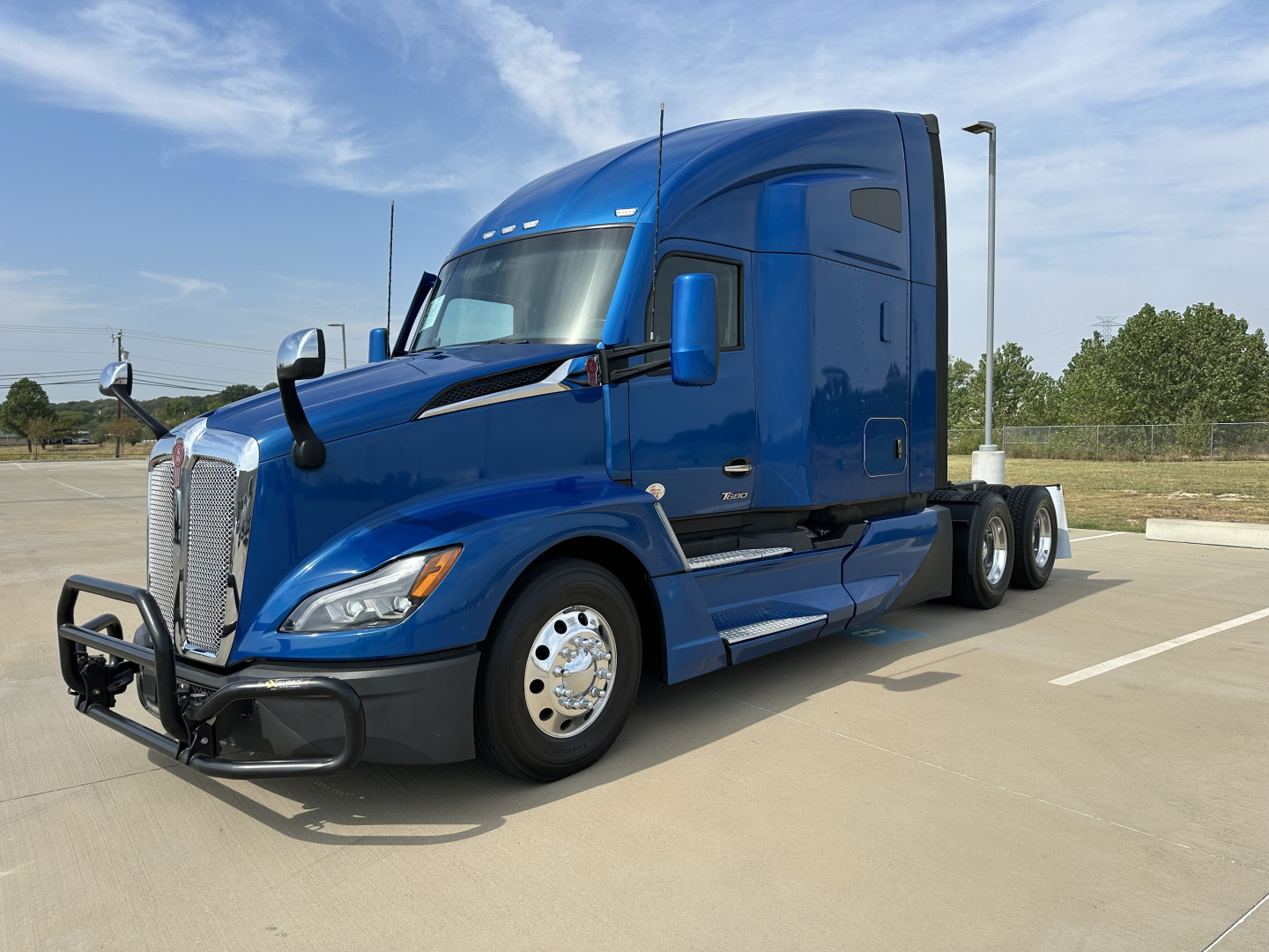 Kenworth T680 tractor-trailer in blue, front-right view, with bumper guard and chrome wheels, highway backdrop.
