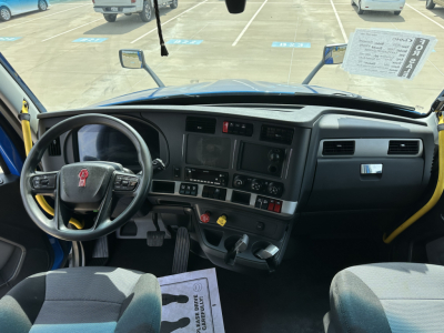 Kenworth T680 cabin dashboard and steering wheel view with controls, pedals, and a window sticker visible; forklift-style yellow grab handles at the doorway.