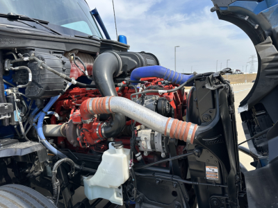 Kenworth T680 engine bay showing coolant reservoir, hoses, and turbo/intercooler pipes. Visible labels and components include a white translucent coolant reservoir, red engine, blue silicone hose, and black radiator shroud.