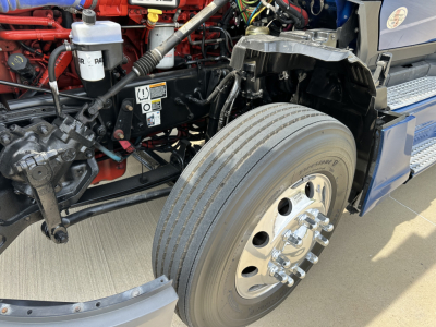 Kenworth T680 front wheel and engine bay detail showing front axle with dual tires and chrome lug nuts, exposed red engine components and hydraulic lines.