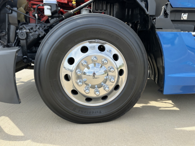Kenworth T680 front wheel and chrome hub showing tire and wheel assembly, engine bay and blue fender in background.