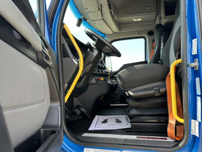 Kenworth T680 truck cab interior showing driver's seat, steering wheel, and dashboard with a yellow guard rail by the door; floor mats visible; blue exterior edge at door.