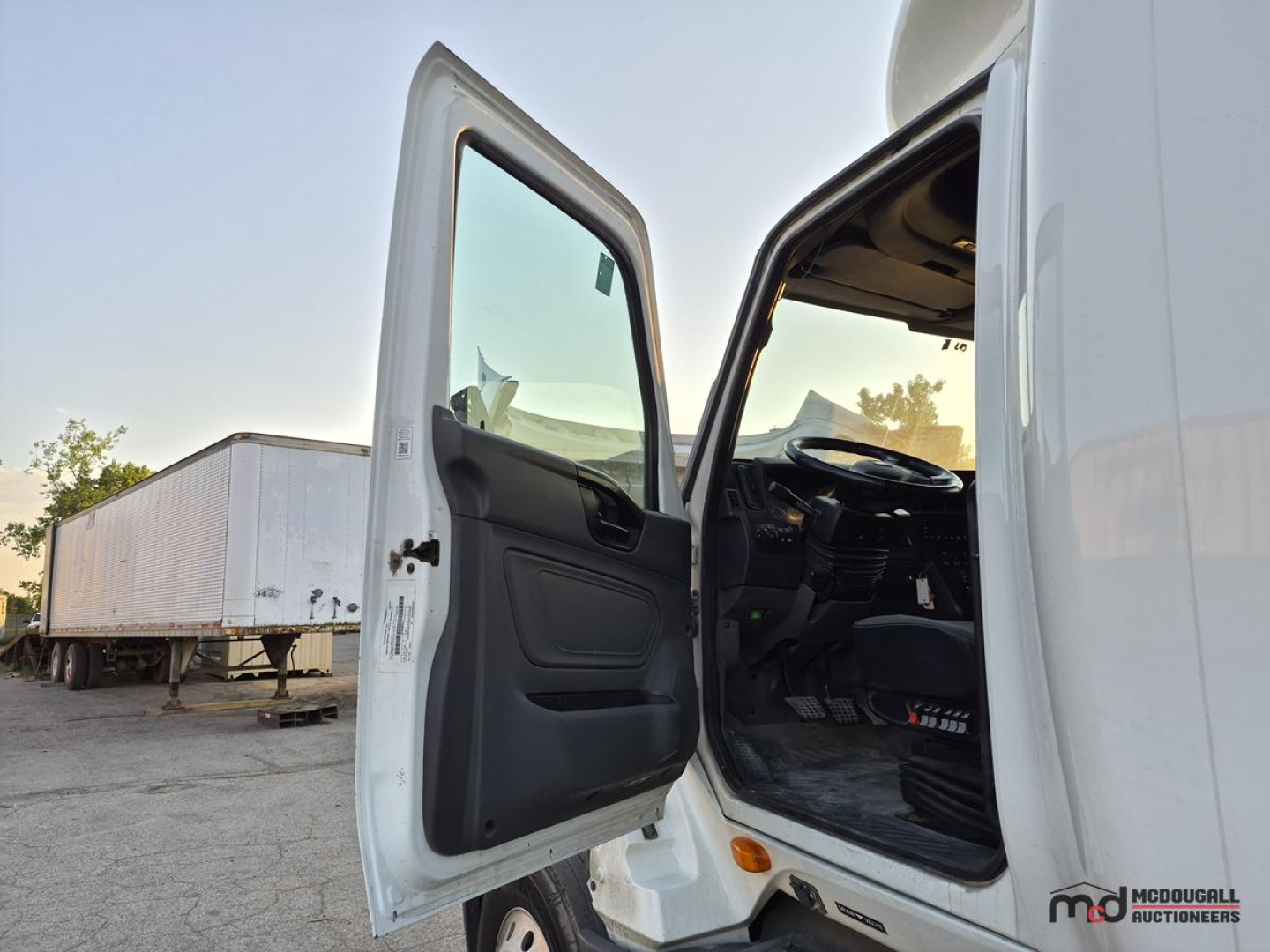 Trucked cab interior visible with driver door open showing dash and steering wheel; exterior white truck with sleeper cab; trailer in background