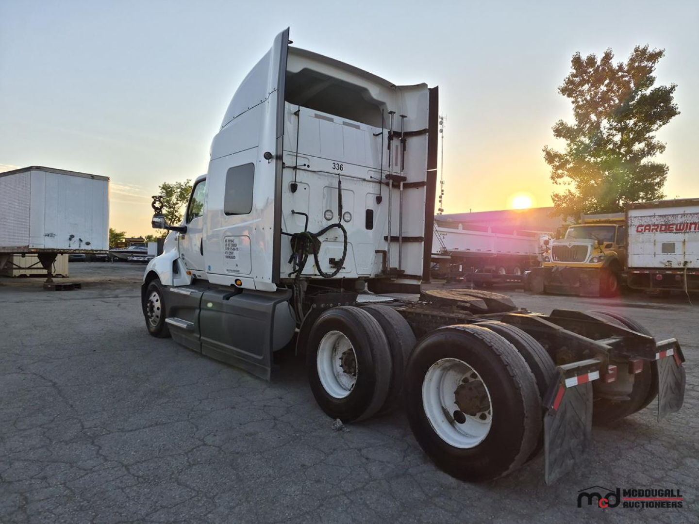 Truck cab and rear view showing fifth wheel and dual rear axles of a sleeper semi, with sunset and trailers in background.