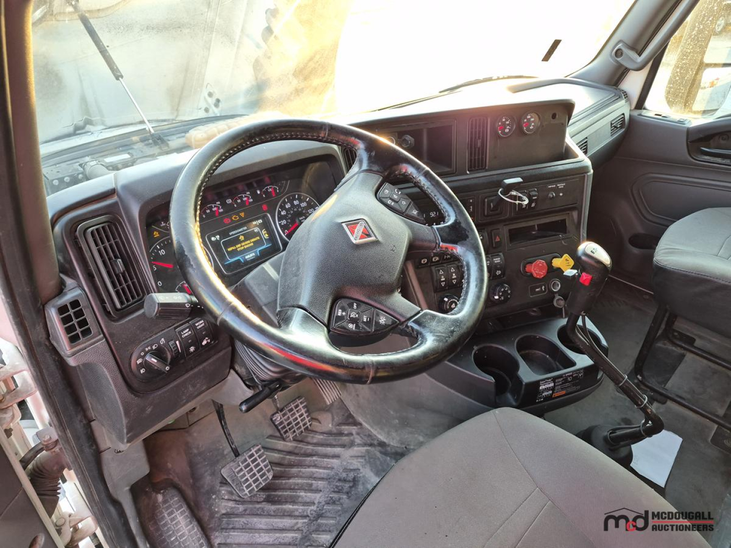Truck cab interior showing steering wheel, instrument cluster, and gear shifter in a sleeper tractor; dashboard features multiple gauges and control switches.