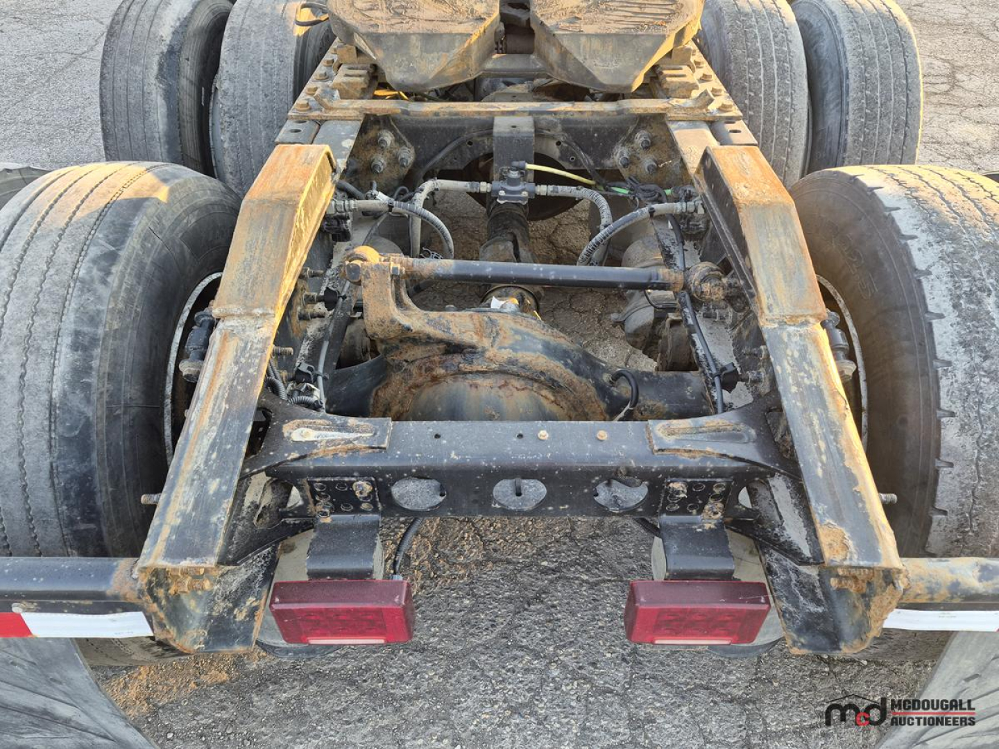 Tractor-trailer chassis view showing rear drive axles and hitch assembly with rusty steel frame and two red taillights.