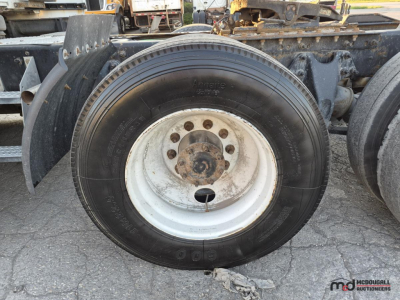 Wheel and tire assembly of a sleeper semi truck showing a large black tire mounted on a white steel wheel with rusted hub, part of a heavy-duty truck chassis