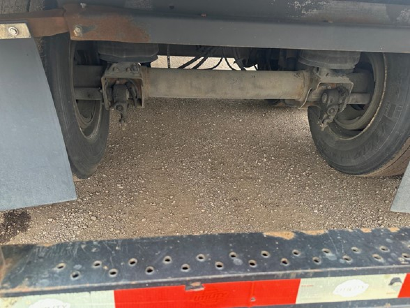 Trailer undercarriage showing tandem axles and differential housing with leaf springs attached, dirt ground beneath.