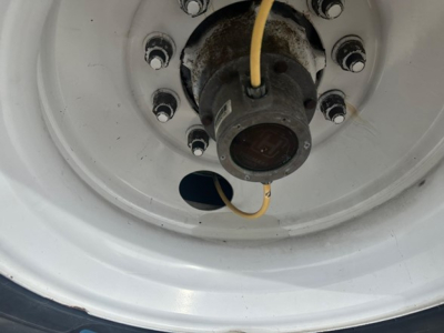 Wheel hub of a trailer with a yellow electrical wire and circular hub cap, white rim, lug nuts around the center, and a dark cylindrical connection at center.