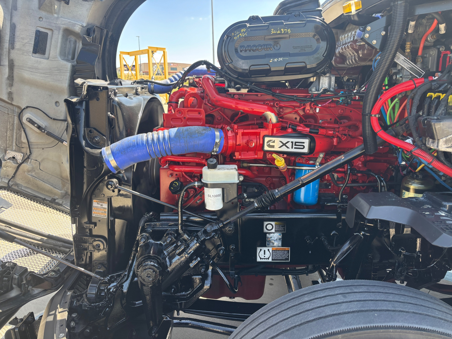 Kenworth T680 engine bay showing red Cummins X15 engine with blue turbo hose, coolant reservoir, and various black hoses and wiring in front engine compartment.