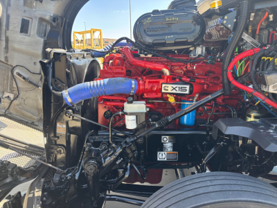 Kenworth T680 engine bay showing red Cummins X15 engine with blue turbo hose, coolant reservoir, and various black hoses and wiring in front engine compartment.