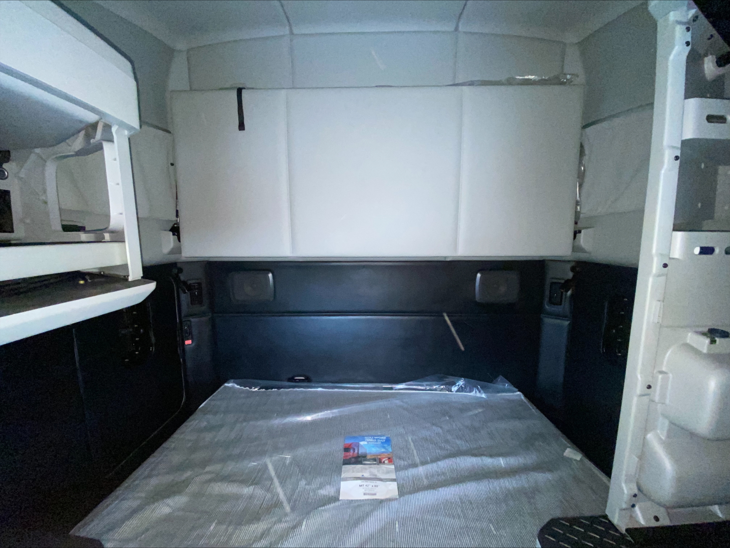Kenworth T680 truck cab interior cargo area with white storage box above and black lower panels; plastic-wrapped mat on floor; small brochure in the center.