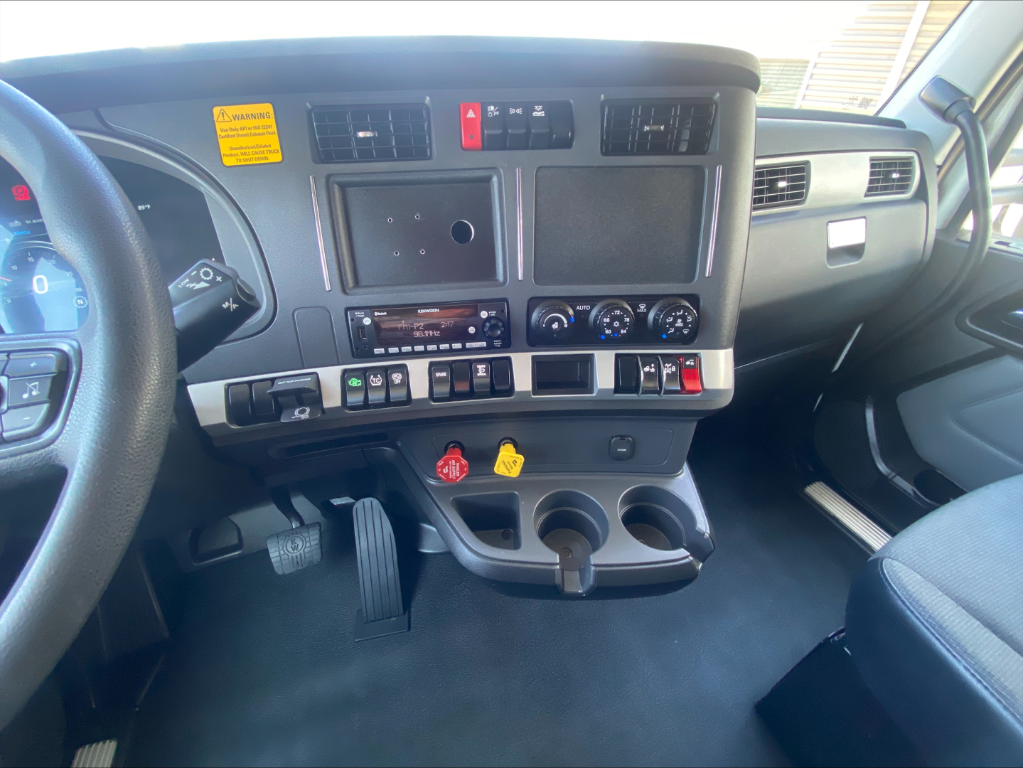 Kenworth T680 cab interior showing dashboard with touchscreen display, control buttons, steering wheel, pedals, and cup holders.