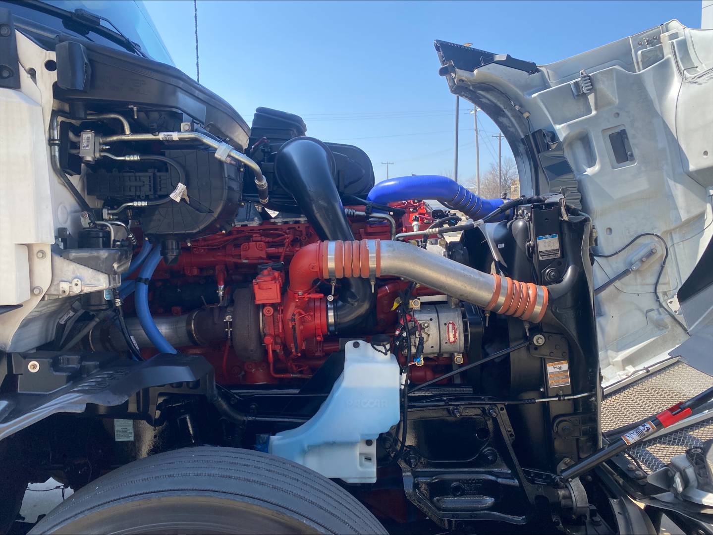 Kenworth T680 engine bay with bright red engine, blue hoses, and white coolant reservoir visible under open hood; multiple components and hoses.