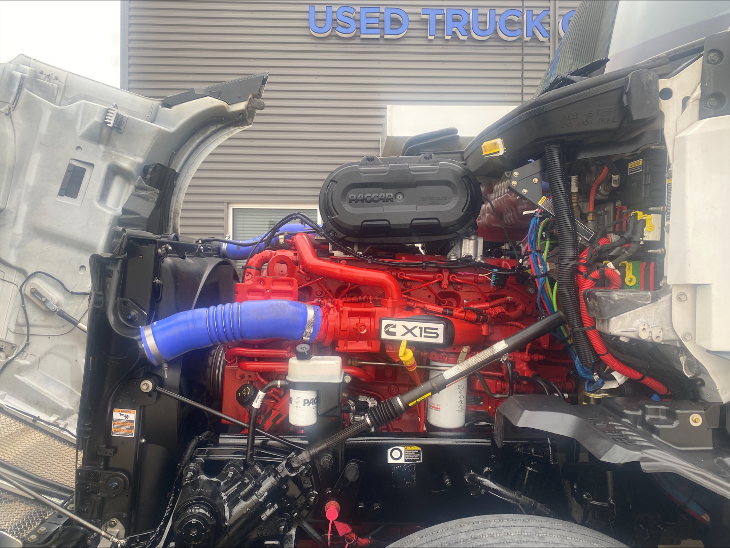 Kenworth T680 engine bay view showing red Cummins X15 engine, blue radiator hose, white coolant reservoir, wiring harnesses, and open hood in front of a truck showroom.
