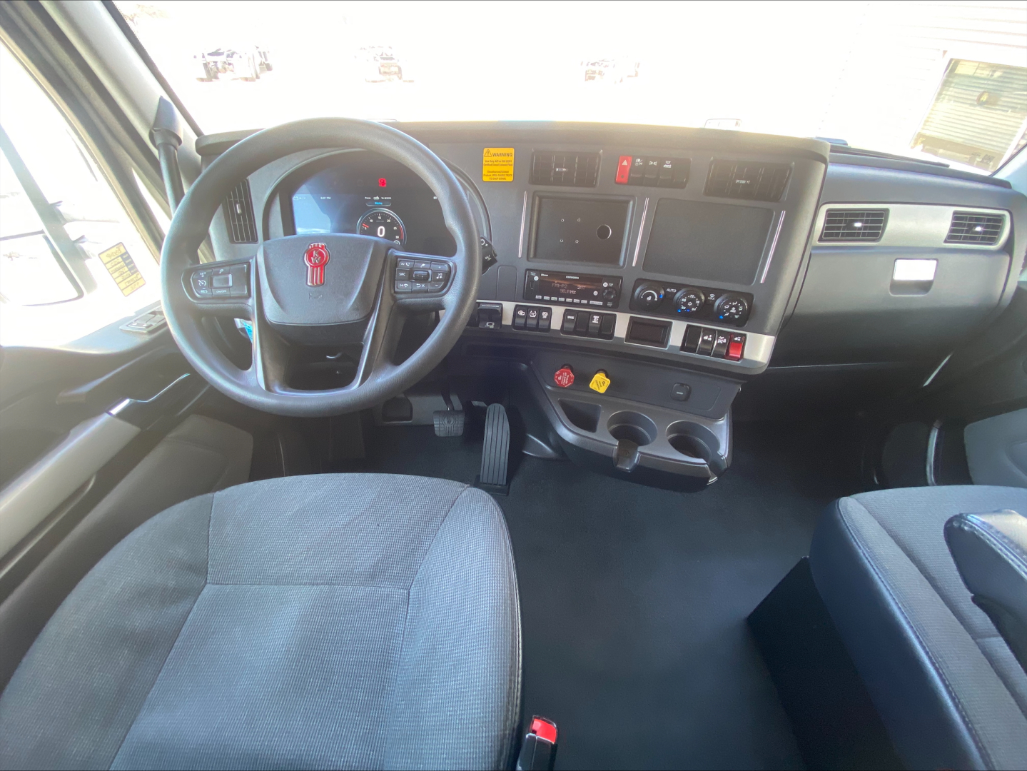 Kenworth T680 cab interior showing steering wheel with red badge, touchscreen display, control panel, and dashboard gauges; center console with climate controls and cup holders.