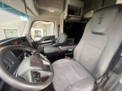 Truck cab interior: steering wheel with Kenworth emblem, driver's seat, dashboard, and passenger seat visible; overhead storage and sleeper area in background.