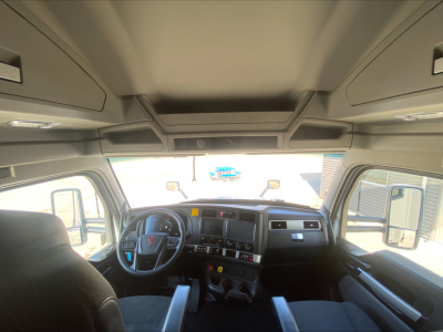 Truck cab interior of 2023 Kenworth T680 showing steering wheel, dashboard, and overhead cabinets. Visible windshield and side windows.