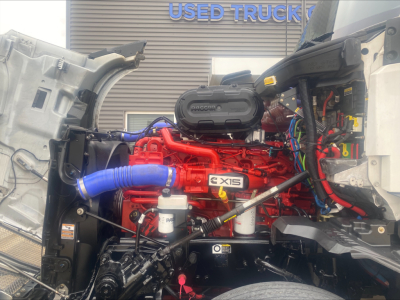 Kenworth T680 engine bay view showing red Cummins X15 engine, blue radiator hose, white coolant reservoir, wiring harnesses, and open hood in front of a truck showroom.