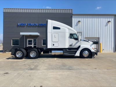 Kenworth T680 side view showing cab with sleeper, triple rear axles, and chrome fuel tanks in front of a blue sky; dealership building in background.