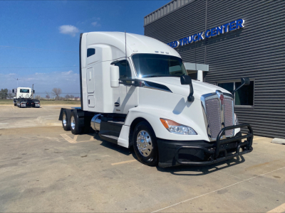 Kenworth T680 conventional sleeper truck in a parking lot, white cab, large grille guard, day cab with chrome mirrors and wheels, factory bumper, dealership building in background.