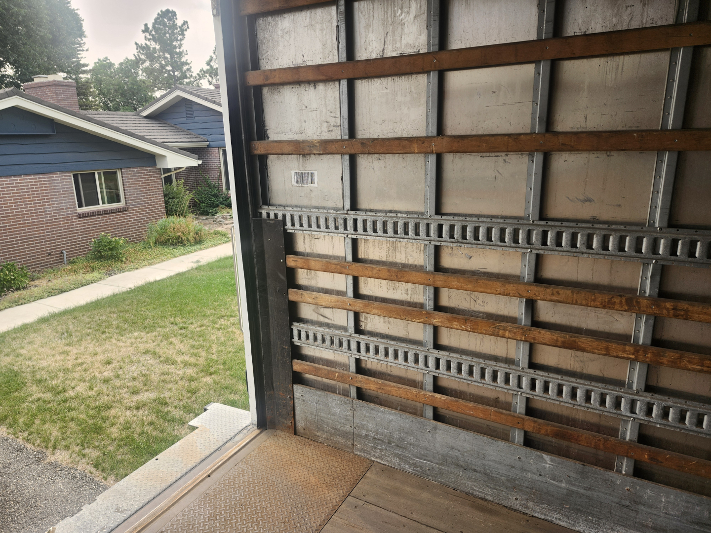 Truck interior wall of a box truck showing metal rails and wooden slats along side, with a small label near the middle. Exterior suburban scene visible through open door.