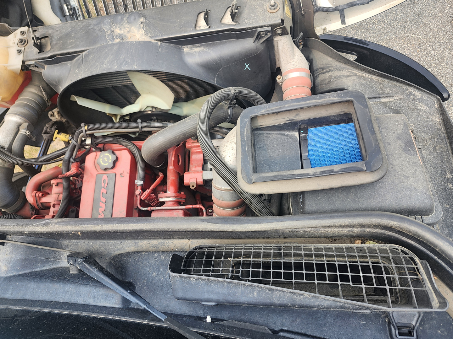 Engine bay of 2020 International MV607 showing Cummins engine with red valve covers, cooling fan, air filter housing, hoses, and radiator grille.