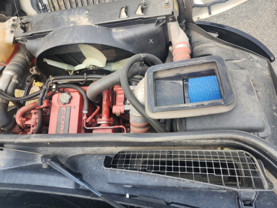 Engine bay of 2020 International MV607 showing Cummins engine with red valve covers, cooling fan, air filter housing, hoses, and radiator grille.