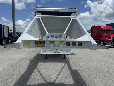 Dump trailer rear view showing raised tailgate with warning labels and stabilizing legs, white metal sides, hitch area and safety decals.