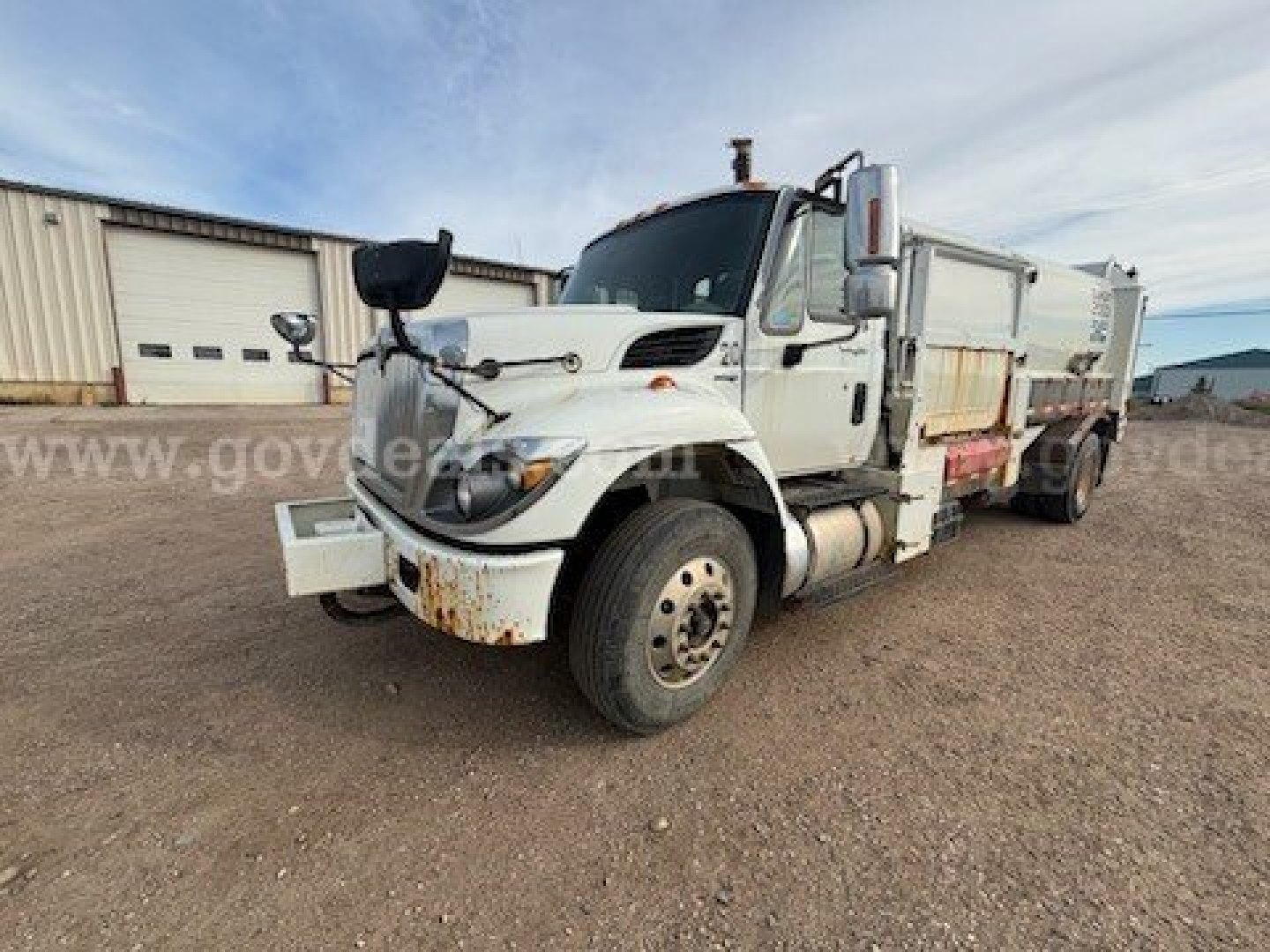 Truck cab and dump body of a white 2011 International 7400 garbage truck with mud flaps and decals, front bumper, and large side mirrors.