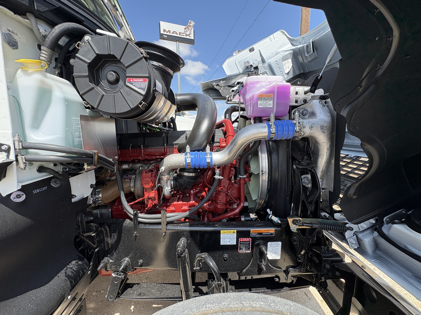Engine bay of a 2027 Mack PI64T conventional daycab showing turbocharger, hoses, coolant reservoir, and engine block with red components.