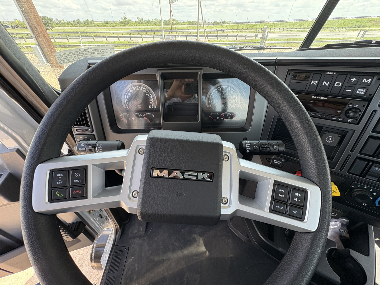 Steering wheel of a Mack truck cab with dashboard gauges and control stalks visible.