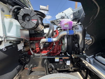 Engine bay of a 2027 Mack PI64T conventional daycab showing turbocharger, hoses, coolant reservoir, and engine block with red components.