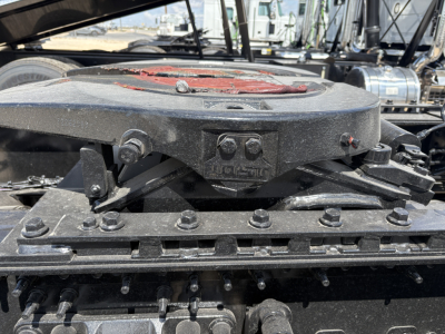 Engine bay area of a Mack truck showing a circular ABS/air reservoir with bolts and mounting plate, with various hoses and frame components visible.