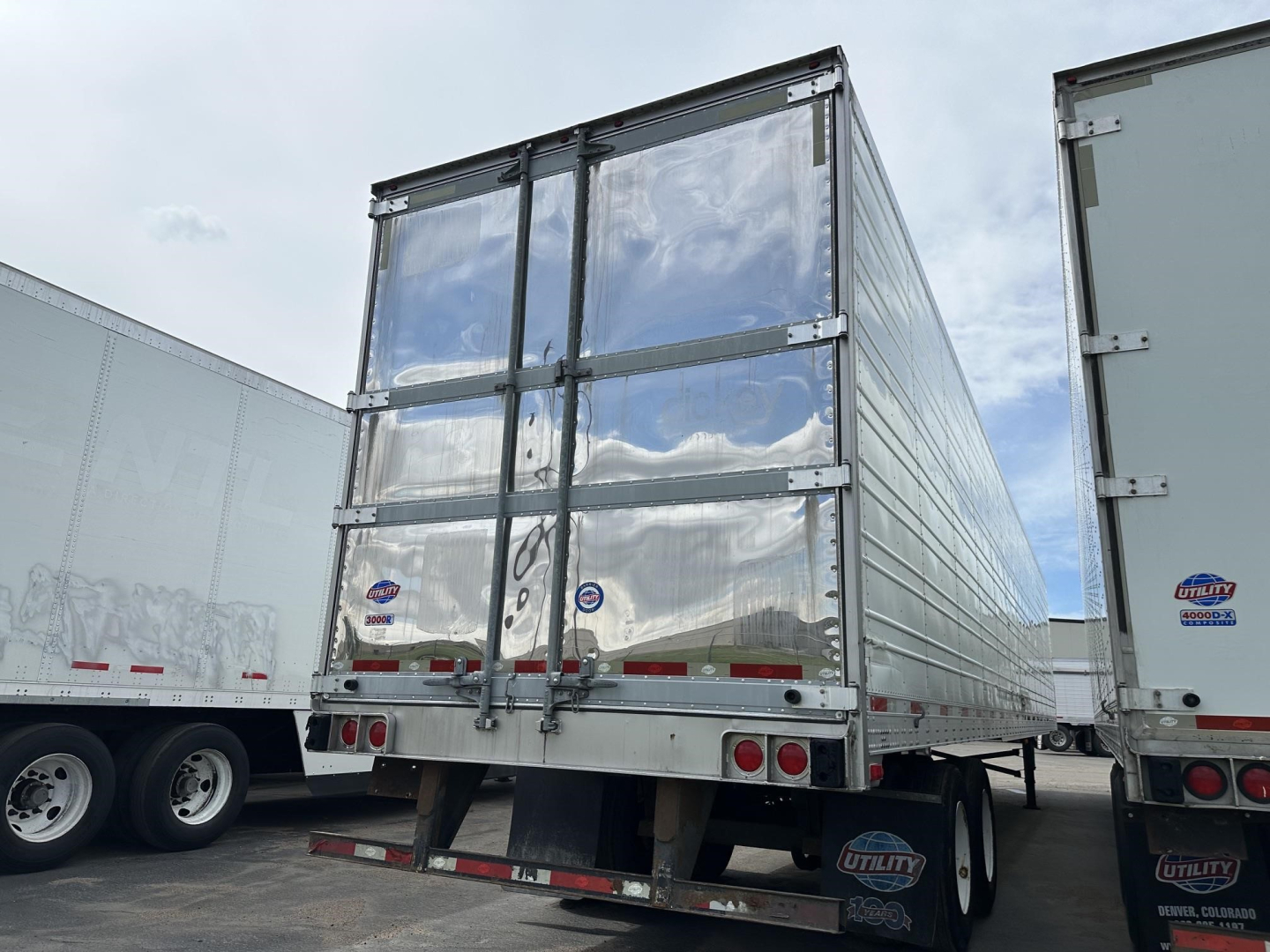 Rear view of a silver Utility 3000R trailer with reflective vertical ribs, dual rear doors, and red taillights. Side of another trailer visible.