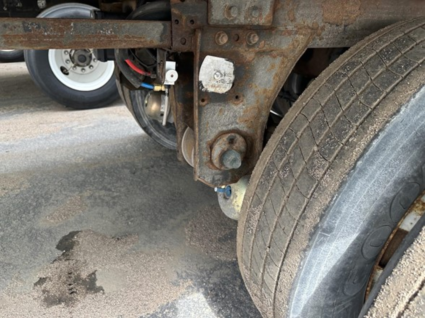 Trailer undercarriage showing rusted frame and dual axles with tires, hydraulic lines, and a white wheel in background.